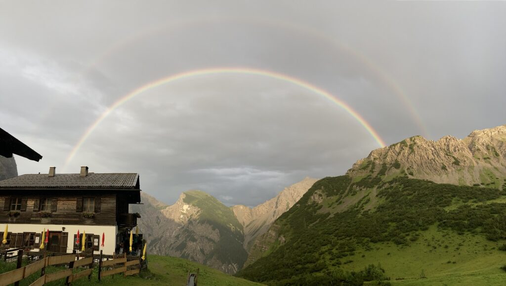 Ein doppelter Regenbogen spannt sich über Berge und ein Holzhaus unter einem wolkenverhangenen Himmel, mit grünen Hügeln im Vordergrund - eine bezaubernde Szene auf der Gramaialm Wanderung.