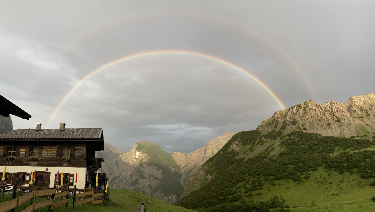 Ein doppelter Regenbogen spannt sich über Berge und ein Holzhaus unter einem wolkenverhangenen Himmel, mit grünen Hügeln im Vordergrund - eine bezaubernde Szene auf der Gramaialm Wanderung.