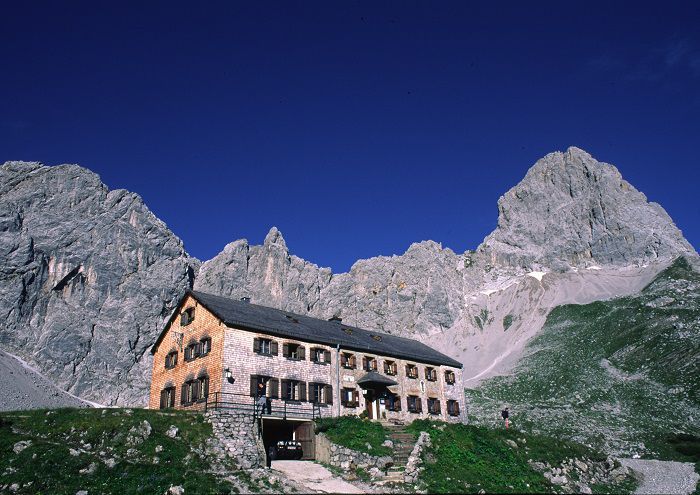 Eine Berghütte aus Stein und Holz steht an einem grasbewachsenen Hang unterhalb der schroffen Gipfel des Lamsenjochs, umgeben von felsigen Bergen und unter einem klaren blauen Himmel.