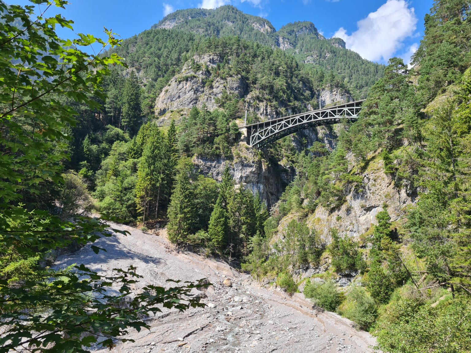 Eine Metallbrücke überspannt eine tiefe, felsige Schlucht entlang der Schlossbachklamm-Wanderung, umgeben von dichtem grünen Wald und Bergen unter einem teilweise bewölkten Himmel.