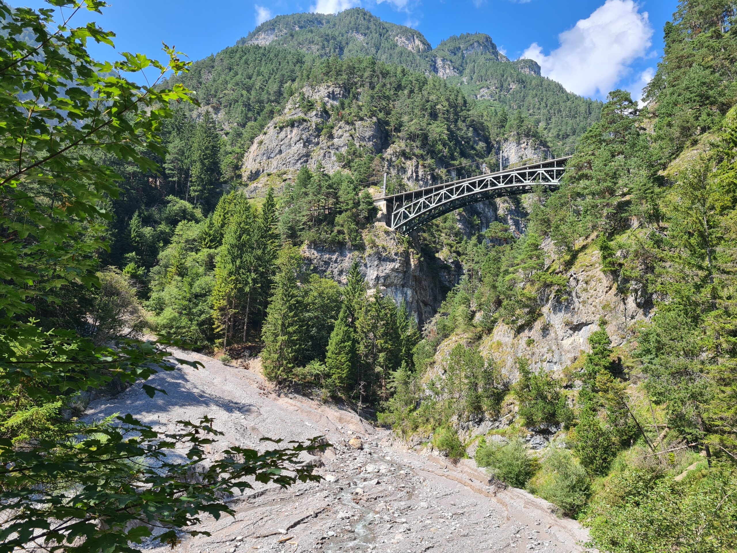 Eine Metallbrücke überspannt eine tiefe, felsige Schlucht entlang der Schlossbachklamm-Wanderung, umgeben von dichtem grünen Wald und Bergen unter einem teilweise bewölkten Himmel.