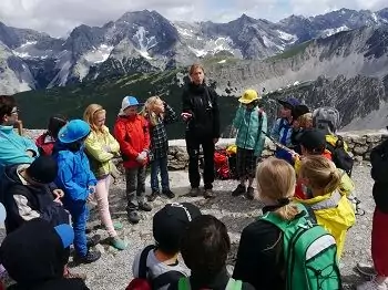 Eine Gruppe von Kindern mit Rucksäcken steht im Freien in einem Kreis und hört einem Erwachsenen zu, während im Hintergrund das majestätische Hafelekar-Gebirge zu sehen ist.