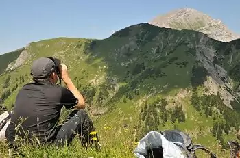 Eine Person sitzt auf einer grasbewachsenen Anhöhe am Großen Ahornboden und blickt durch ein Fernglas auf einen fernen Berg unter klarem Himmel, in der Nähe ein Rucksack.