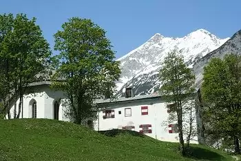 Ein weißes Gebäude und eine kleine Kapelle stehen auf einem grasbewachsenen Hügel mit Bäumen im Halltal, umgeben von schneebedeckten Bergen und unter einem klaren blauen Himmel.