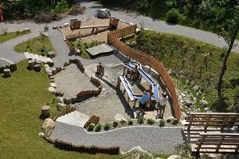 Luftaufnahme eines kleinen Spielplatzes im Freien in Halltal mit einem Wasserspielbereich, einem Sandkasten, Holzstrukturen, Picknicktischen sowie Erwachsenen und Kindern, die auf dem Platz interagieren.