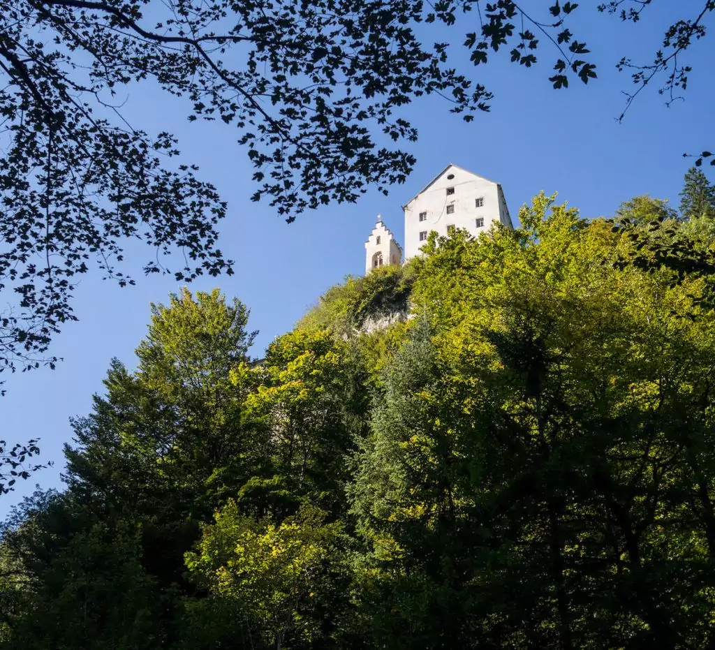 Ein weißes Schloss steht auf einem bewaldeten Hügel, teilweise von grünen Bäumen verdeckt, mit einem klaren blauen Himmel im Hintergrund, der an die Aussicht entlang des Wolfsklammweges erinnert.