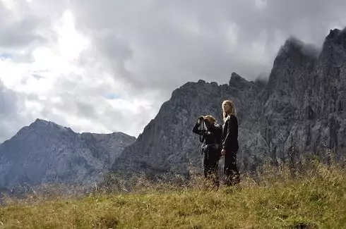Zwei Menschen stehen auf einer Wiese in der Nähe der zerklüfteten Lalidererwände unter einem wolkenverhangenen Himmel; einer schaut durch ein Fernglas, der andere blickt nach vorn.