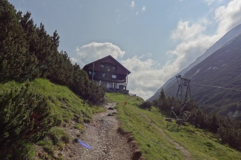 Ein Schotterweg, Teil des Tiroler Adlerwegs, führt bergauf zu einer hölzernen Berghütte, umgeben von grünen Büschen und grasbewachsenen Hängen unter einem teilweise bewölkten Himmel.