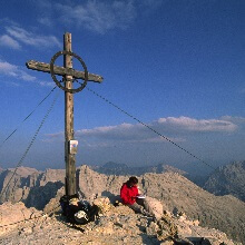Eine Person sitzt bei klarem Himmel neben einem großen, mit Seilen befestigten Holzkreuz auf einem felsigen Berggipfel am berühmten Tiroler Adlerweg.
