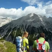 Zwei Wanderer mit Rucksäcken stehen an einem grasbewachsenen Hang und blicken auf eine Bergkette unter einem teilweise bewölkten Himmel - und fangen damit den Geist von München Venedig Weitwandern ein.