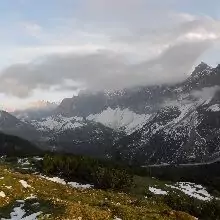 Die schneebedeckten Berge der Via Alpina Karwendel sind teilweise von Wolken bedeckt, im Vordergrund liegen Schneeflecken auf grasbewachsenem Gelände bei bewölktem Himmel.