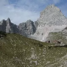 Ein grasbewachsener Hang mit verstreuten Felsen im Vordergrund und hohe, schroffe Berge unter einem teilweise bewölkten Himmel bilden eine klassische Szene entlang der Via Alpina Karwendel.