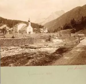 Sepiafarbenes Foto von Hinterriß, einem kleinen Dorf mit einer Kirche, Holzzäunen, einem Feldweg und Bergen im Hintergrund.