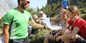 Ein Mann in einem grünen Hemd zeigt einer Gruppe von Menschen, die im Freien auf einem felsigen Gelände in der Nähe von Hinterriß sitzen, eine Landkarte, mit Bergen und Bäumen im Hintergrund.