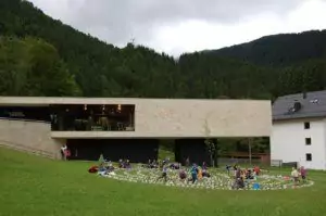 Modernes Gebäude mit großen Fenstern neben einem weißen Haus in Hinterriß, auf einer Wiese, auf der sich Menschen in einem Kreis versammeln, umgeben von bewaldeten Hügeln.