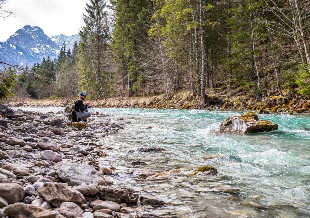 Person, die neben einem felsigen Flussufer in einem Waldgebiet mit Bergen im Hintergrund hockt und die nachhaltige Fischerei am Rissbach beobachtet, während der Fluss unter einem teilweise bewölkten Himmel schnell fließt.