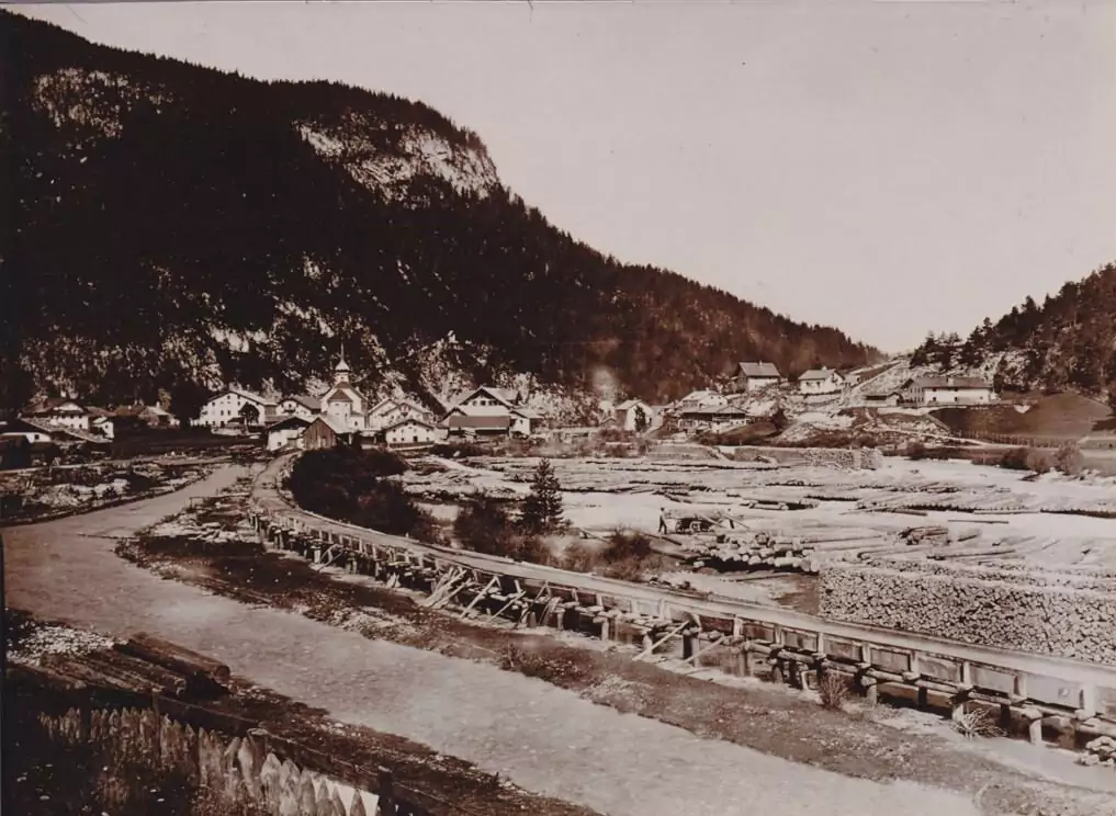 Ein kleines Dorf mit Holzhäusern liegt am Fuße eines bewaldeten Berges, mit einer Brücke und einem Flussbett im Vordergrund - eine ideale Kulisse für ein Interregprojekt, das sich auf grenzüberschreitende Zusammenarbeit und Gemeindeentwicklung konzentriert.