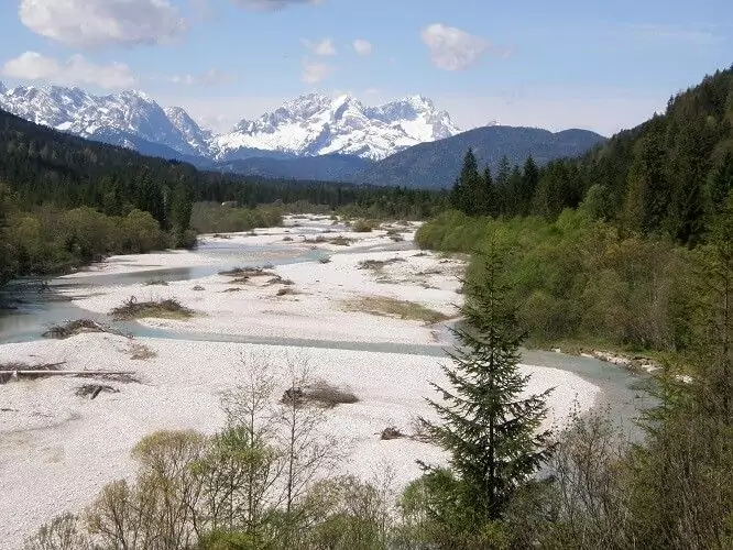 Ein Fluss mit Kiesbänken fließt durch ein bewaldetes Tal, das Teil eines Interregprojekts ist. Im Hintergrund sind schneebedeckte Berge unter einem teilweise bewölkten Himmel zu sehen.