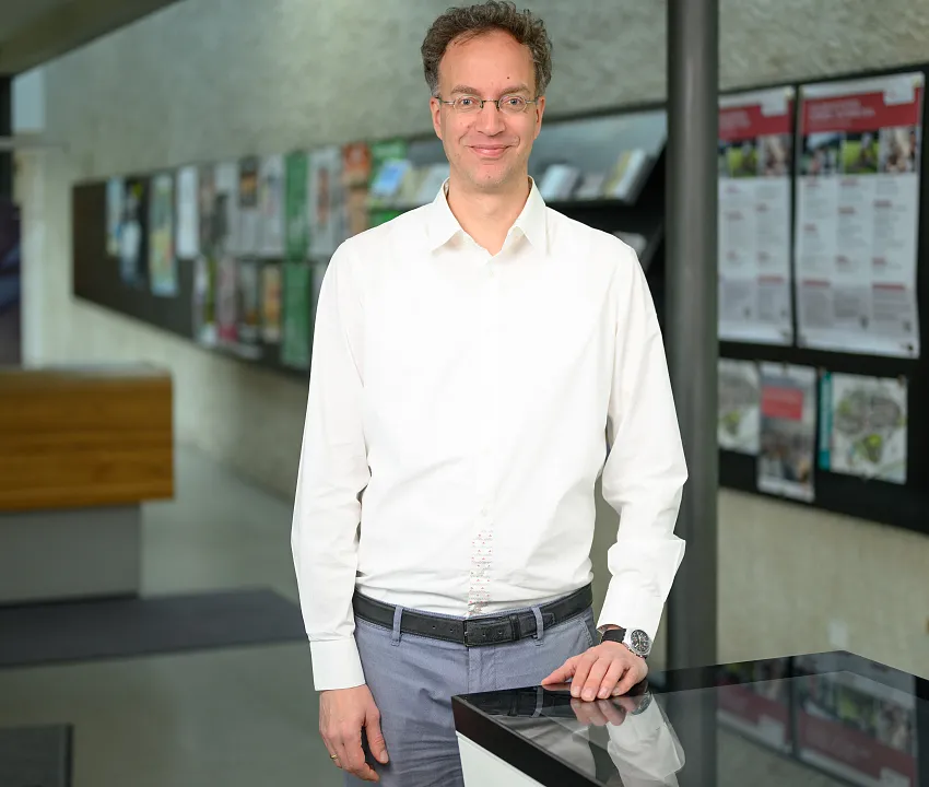 Martin Friede, gekleidet in ein weißes Hemd und eine graue Hose, steht drinnen an einem Tresen, an der Wand hinter ihm sind Broschüren und Informationsschilder zu sehen.