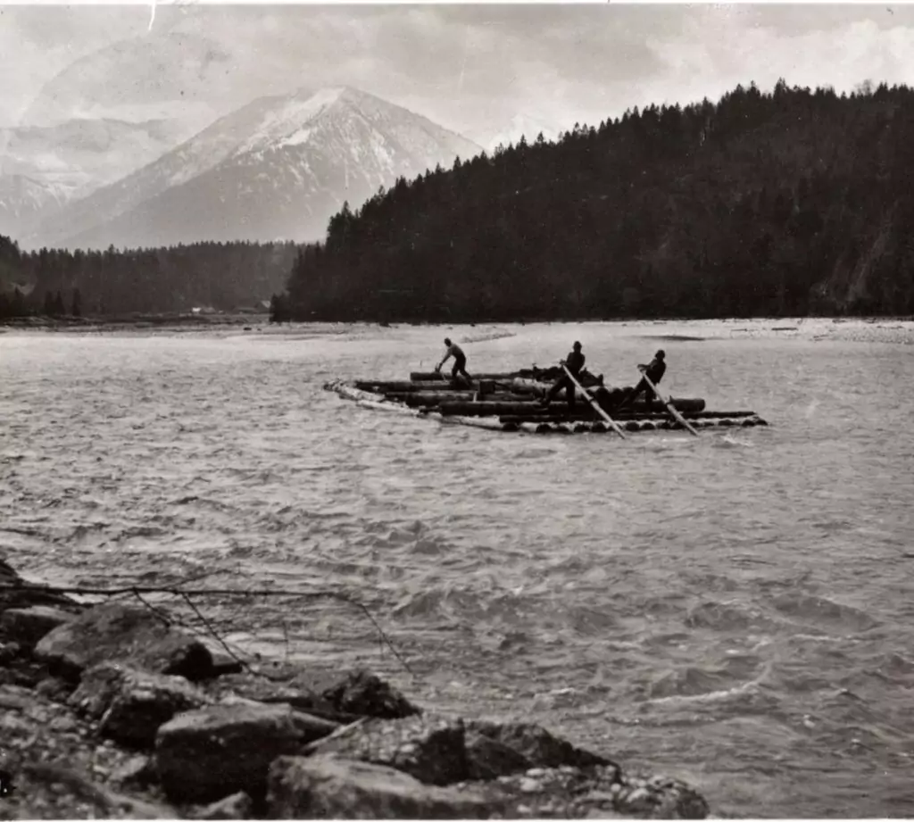 Vier Personen fahren auf einem Holzfloß im traditionellen Flößerstil einen Fluss hinunter, umgeben von dichten Wäldern und schneebedeckten Bergen im Hintergrund.