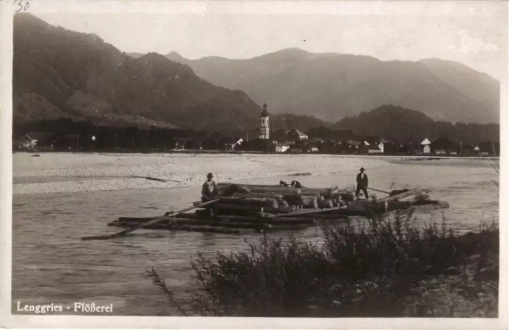 Schwarz-Weiß-Foto von drei Personen, die auf einem großen Holzfloß stehen und die traditionelle Flößerei auf einem Fluss aufnehmen, mit einem Dorf und Bergen im Hintergrund. Die Bildunterschrift lautet "Lenggries - Flößerei".