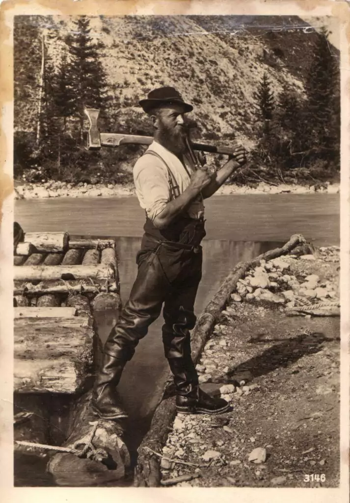 Ein bärtiger Mann steht an einem Fluss, hält eine Axt über der Schulter, trägt hohe Stiefel, Hosenträger und einen Hut, der an die traditionellen Flößer erinnert, und ist von Baumstämmen und Bäumen umgeben.