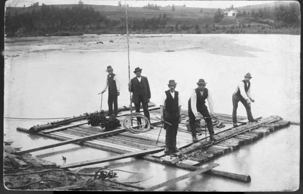 Fünf Männer in förmlicher Kleidung stehen auf einem großen Holzfloß, das auf einem Fluss schwimmt, und demonstrieren die traditionelle Flößerei, während sie Werkzeuge und Seile halten; im Hintergrund ist eine ländliche Landschaft zu sehen.