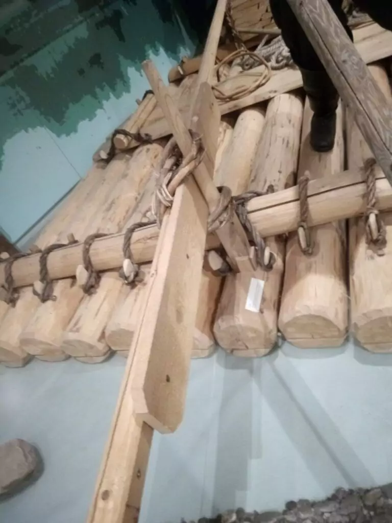 Nahaufnahme eines Holzfloßes, das in der Tradition der Flößerei aus mit Seilen zusammengehaltenen Baumstämmen und einem einfachen hölzernen Steuermechanismus gefertigt wurde.