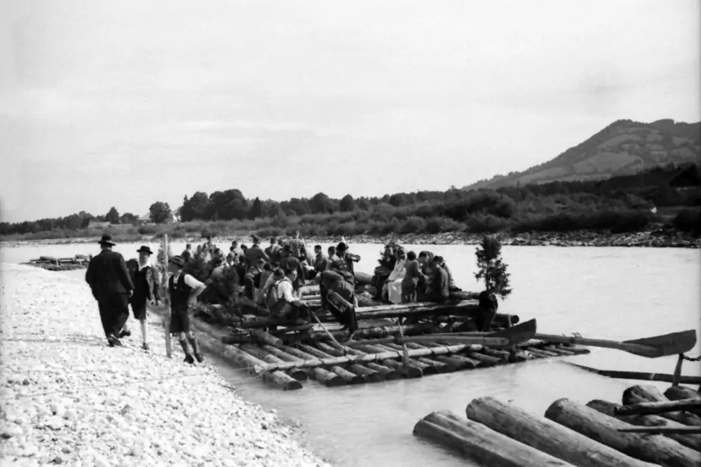Eine Gruppe von Menschen steht und sitzt auf großen Holzflößen am Flussufer und übt sich in traditioneller Flößerei, wobei im Hintergrund Berge und Bäume zu sehen sind.