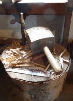 Ein Holzhammer und ein Meißel liegen auf einem Baumstumpf, umgeben von Holzspänen und einem Schild mit der Aufschrift "Hechenzahnschläger", das auf die Kunst der Bearbeitung von Holz als Werkstoff hinweist.
