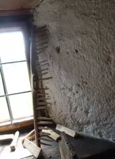 Ein Holzrechen aus robustem Werkstoff Holz lehnt an einer rauen, weiß verputzten Wand neben einem Fenster, unter dem verstreute Holzbretter liegen.