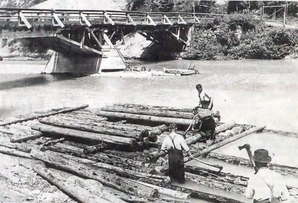 Vier Personen stehen auf schwimmenden Baumstämmen und üben sich in Flößerei, während sie ein Holzfloß auf einem Fluss in der Nähe einer Holzbrücke zusammenbauen.