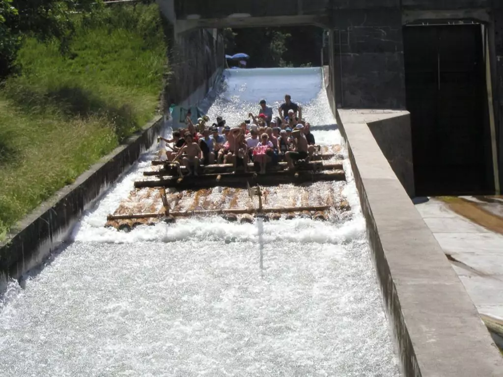 Eine Gruppe von Menschen genießt die Flößerei, während sie auf einem großen Holzfloß eine Wasserrutsche aus Beton hinunterfährt, die von Grünflächen und Steinmauern umgeben ist.