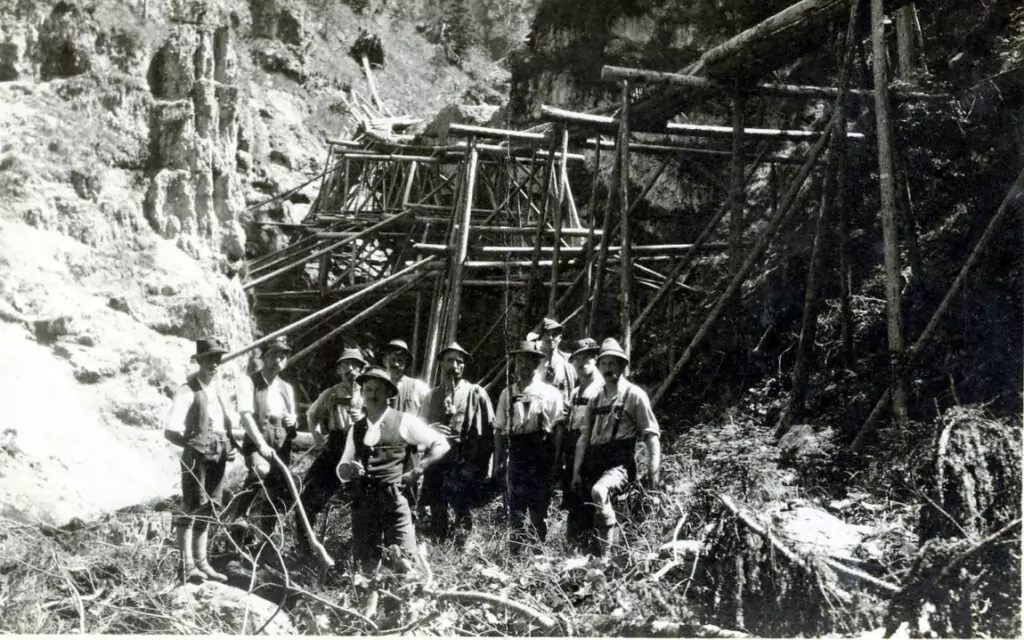 Eine Gruppe von Männern mit Hüten steht vor einer großen Holzkonstruktion namens Loiten und Riesen, die in einen felsigen, bewaldeten Hang gebaut ist.