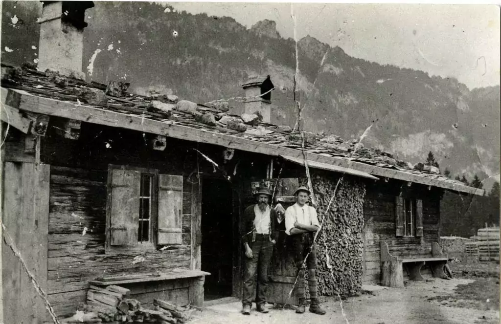 Zwei Männer stehen vor einer rustikalen Kobel-Holzhütte, deren steingedecktes Dach und gestapeltes Brennholz sich perfekt in den Hüttencharme der Bergkulisse einfügt.