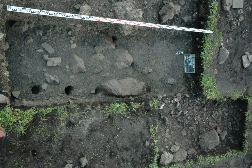 Luftaufnahme einer archäologischen Ausgrabungsstätte in der Nähe von Saline Hall, mit Steinen, Bodenschichten, Messband und einer kleinen Informationstafel.