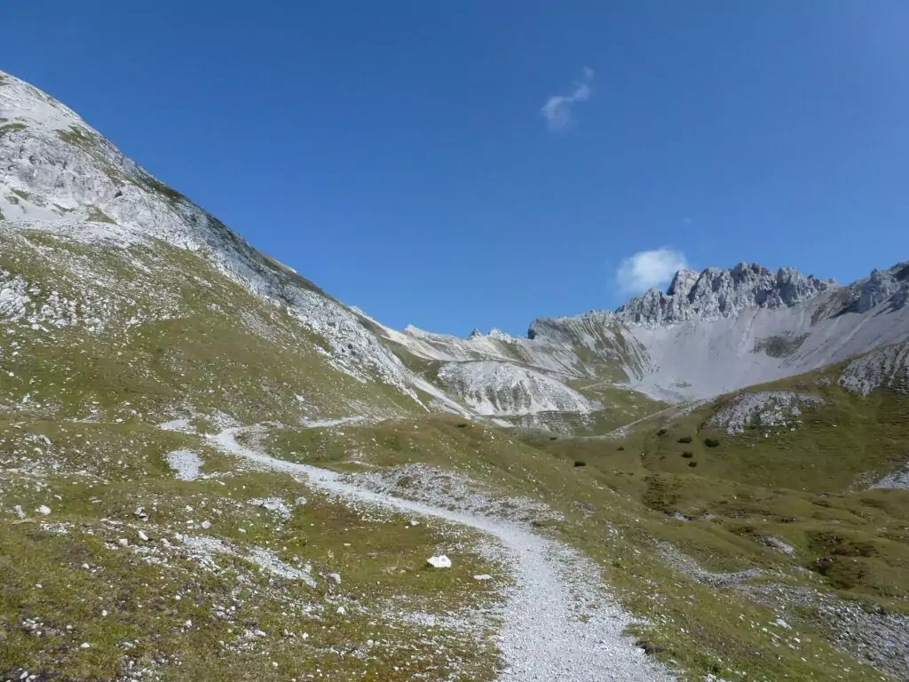 Ein kurvenreicher Schotterweg führt durch ein grasbewachsenes Bergtal mit felsigen Hängen unter einem strahlend blauen Himmel.
