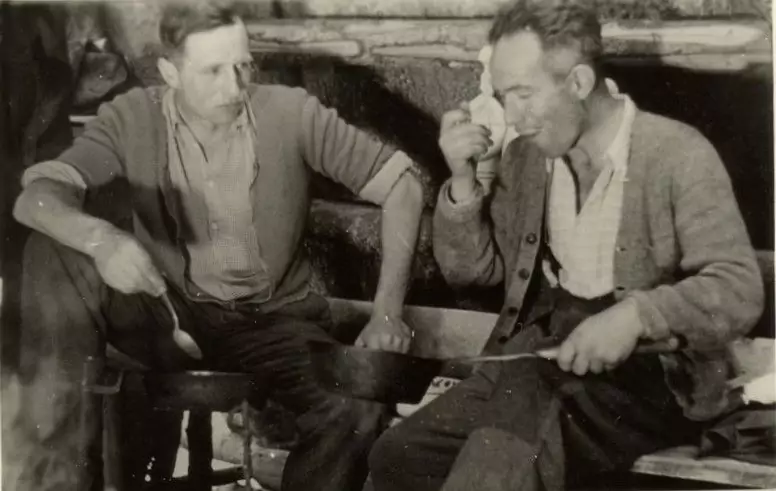 Zwei Männer sitzen in einem Haus; der eine isst aus einem Löffel und hält eine Schüssel in der Hand, der andere hält eine Pfanne und einen Löffel, beide sind leger gekleidet.