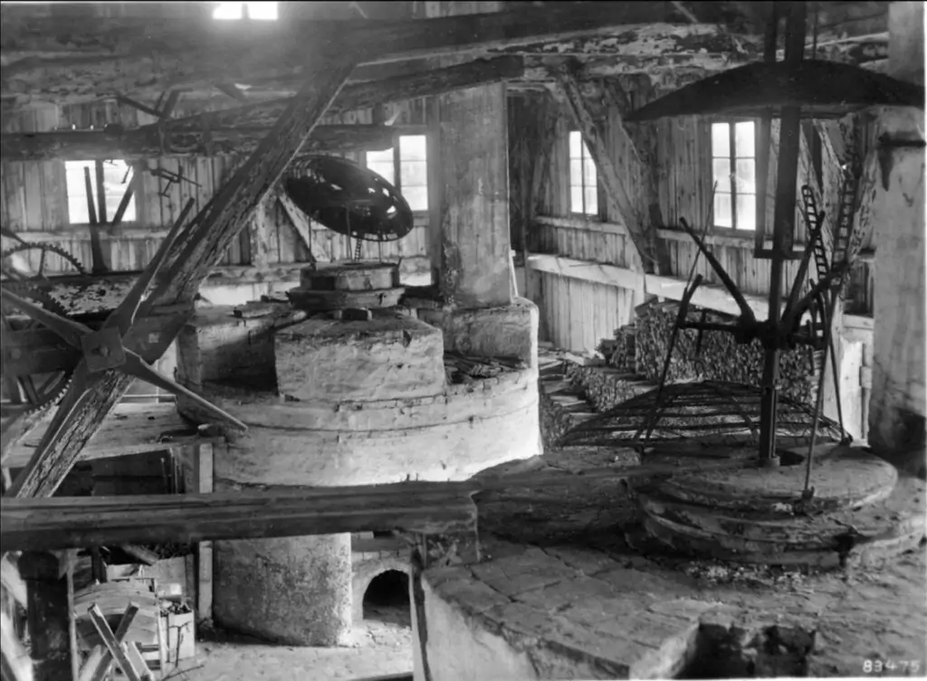Schwarz-Weiß-Foto des Innenraums einer alten Mühle mit großen steinernen Mühlrädern, Holzbalken, Maschinen und Fenstern, die Tageslicht hereinlassen.