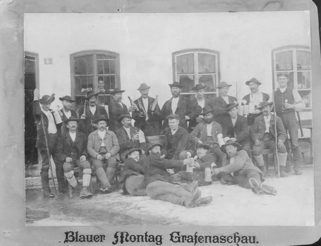 Eine Gruppe von Männern in traditioneller Kleidung posiert vor einem Gebäude, einige sitzend, einige stehend, mit Bierkrügen und Flaschen. Der Text darunter lautet: "Blauer Montag Grafenaschau.