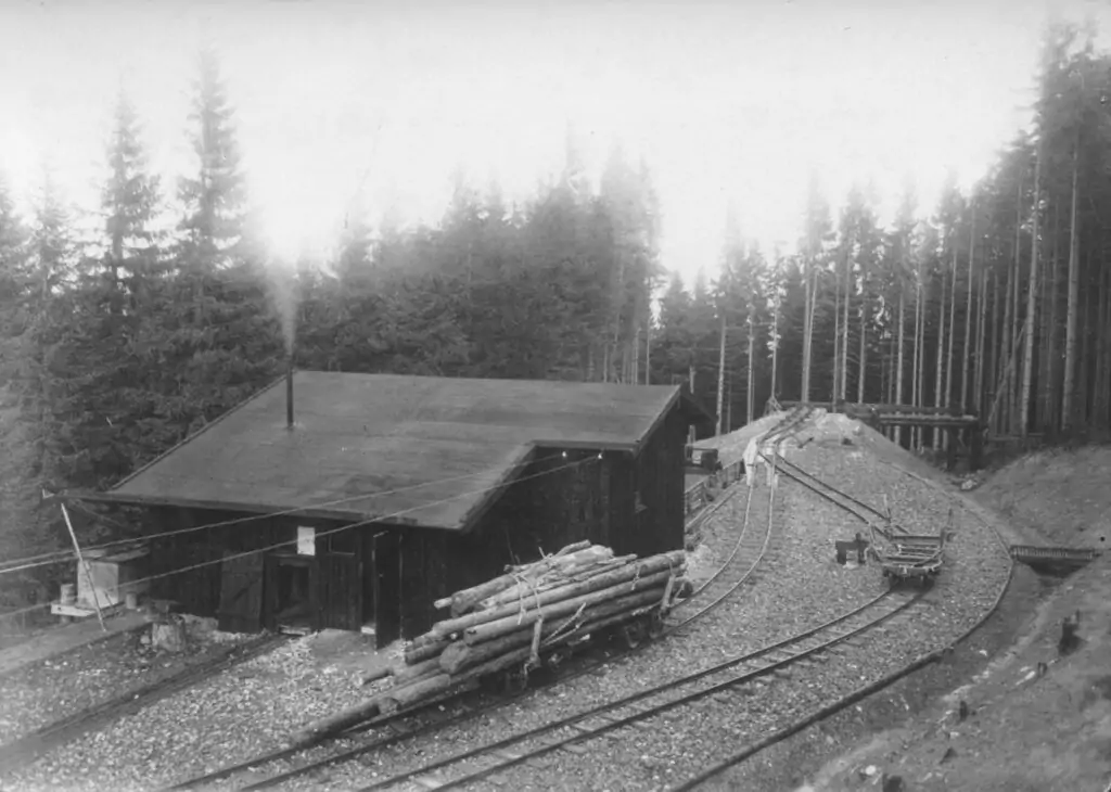 Schwarz-Weiß-Foto eines kleinen Holzgebäudes an Eisenbahnschienen, mit gestapelten Baumstämmen in der Nähe und der Bremsbergbahn, die durch einen dichten Wald im Hintergrund verläuft.