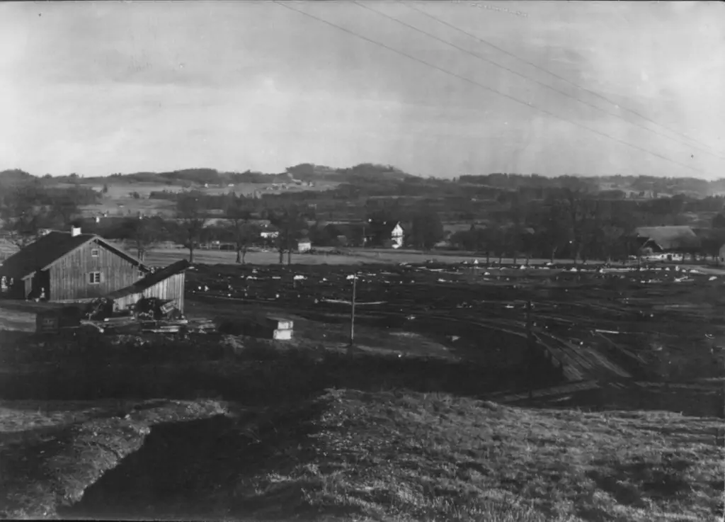Schwarz-Weiß-Foto einer ländlichen Landschaft mit einem Holzgebäude im Vordergrund, offenen Feldern, verstreuten Bäumen und entfernten landwirtschaftlichen Gebäuden unter einem bewölkten Himmel - die Gleise der Bremsbergbahn sind in der Ferne schwach zu erkennen.
