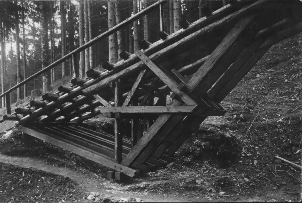 Eine Holzkonstruktion mit Balken und Schienen an einem bewaldeten Hang, möglicherweise Teil eines alten Bremsbergbahn-Bergbau- oder Holzfällerbetriebs, mit Bäumen im Hintergrund.
