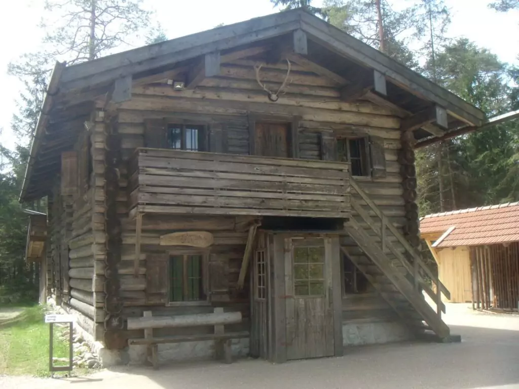 Ein zweistöckiges Holzblockhaus mit Balkon, Außentreppe und einem Schild über der Tür, das in einem Waldgebiet in der Nähe des Holzknechtmuseums Ruhpolding liegt.