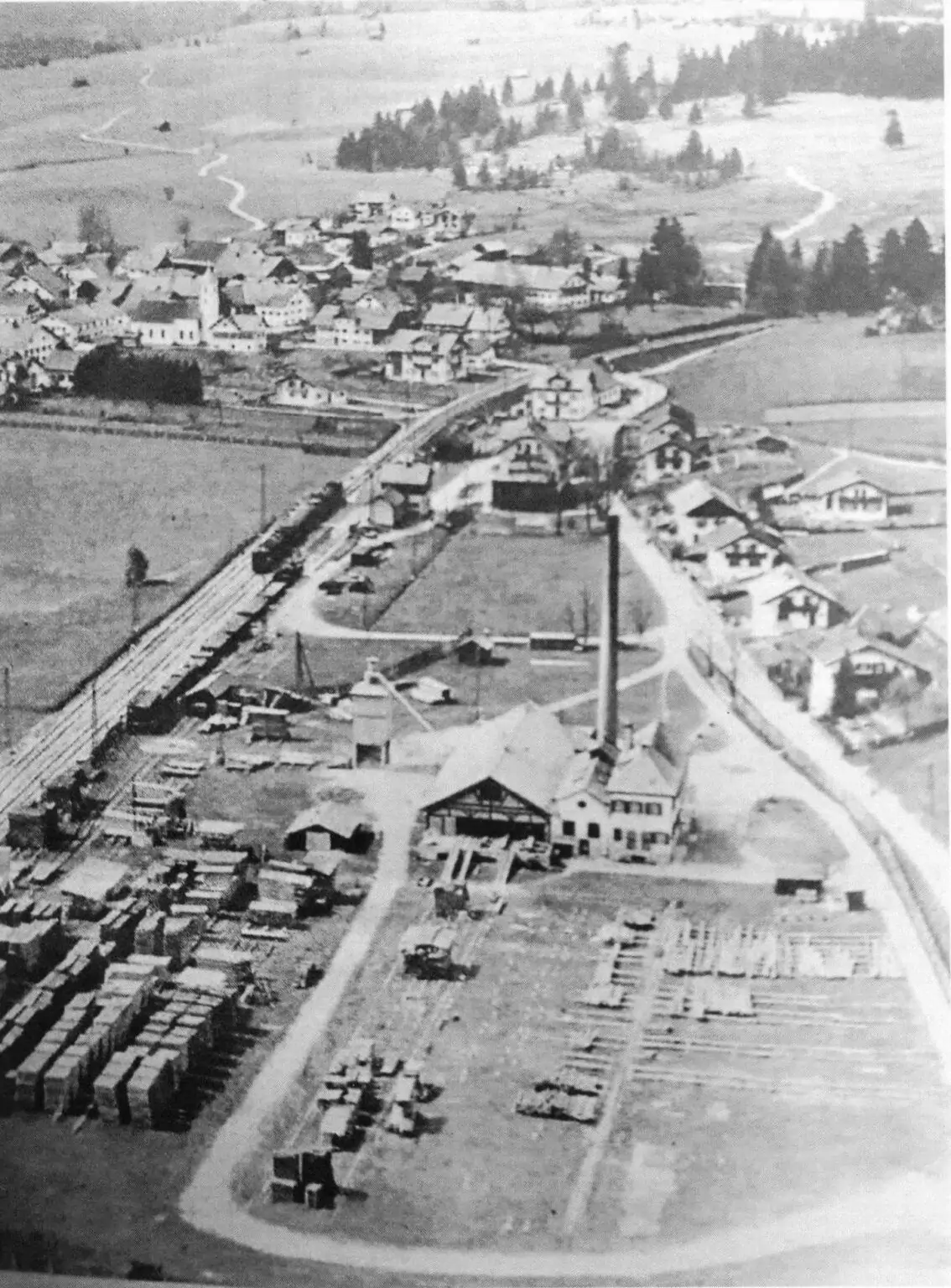 Schwarz-Weiß-Luftaufnahme eines ländlichen Dorfes mit Häusern, einer großen Industrieanlage und ordentlich gestapelten Holzstapeln im Vordergrund, umgeben von Feldern und Bäumen.