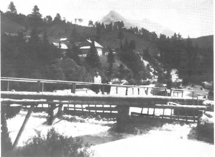Schwarz-Weiß-Foto einer Holzbrücke über einen Fluss, auf der eine Person steht, umgeben von Bäumen und Gebäuden, mit einem Berg im Hintergrund.