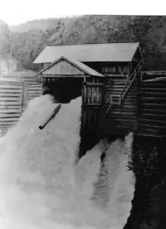 Schwarz-Weiß-Foto einer Wasserrutsche, die Wasser und einen Baumstamm aus einer Holzkonstruktion abwirft, mit bewaldeten Hügeln im Hintergrund.