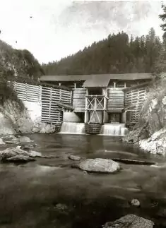 Schwarz-Weiß-Foto einer hölzernen Dammkonstruktion, die einen Fluss überspannt, mit bewaldeten Hügeln im Hintergrund.