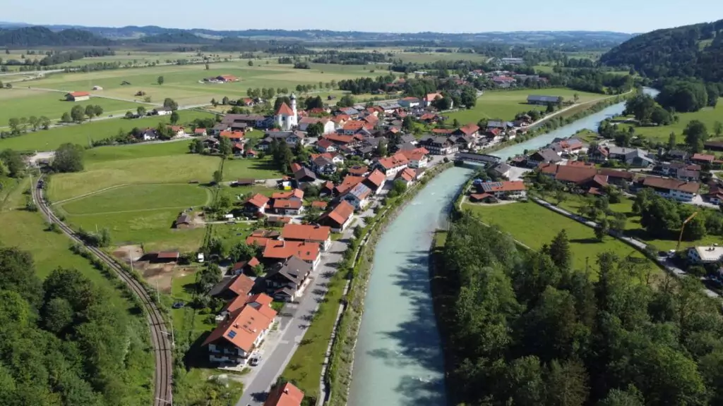 Luftaufnahme eines kleinen Dorfes mit roten Dächern entlang eines gewundenen Flusses, umgeben von grünen Feldern und fernen Hügeln.
