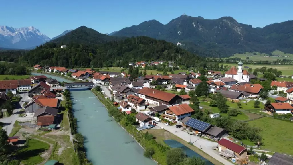 Luftaufnahme eines kleinen Dorfes mit roten Dächern, einem Fluss, einer Brücke und Bergen im Hintergrund unter einem klaren blauen Himmel.